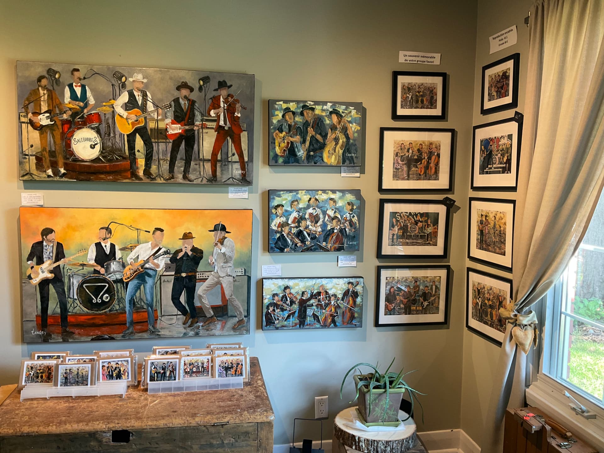 Mur de la galerie-atelier couvert de toiles de musiciens et de reproductions encadrées, cartes d'art posées sur une table de bois.